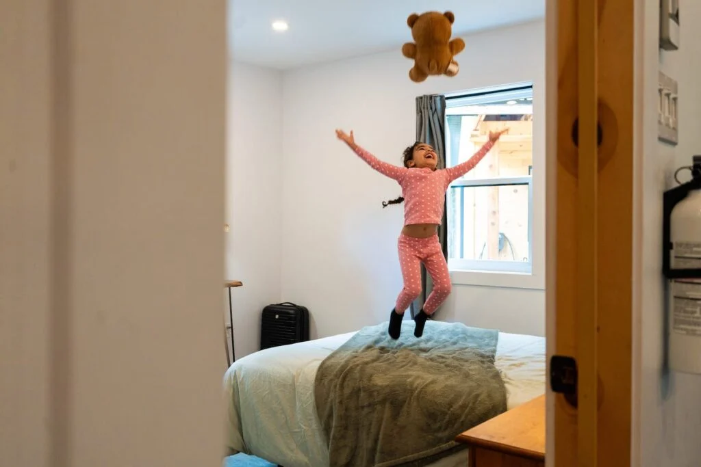 Une fillette en pyjama saute de joie sur le lit en lançant son ourson, dans une chambre d’un chalet Astra